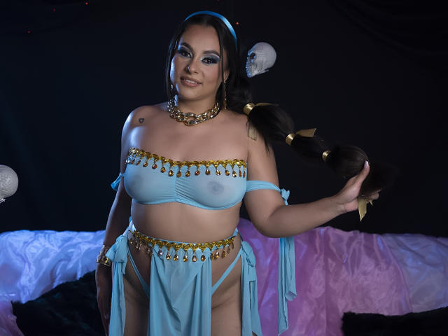 LunaSoler, avec de longs cheveux bruns, un bandeau bleu, un haut bleu transparent qui révèle ses seins, un collier en chaîne dorée et une jupe assortie, se tient debout, souriante, tout en se tenant les cheveux, et portant des bracelets dorés..