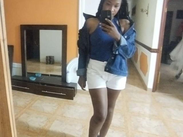 TaniaDoux with dark skin, wearing a blue denim jacket and white shorts, showing her bare ass in a mirror selfie, holding a phone, standing in a tiled room.
