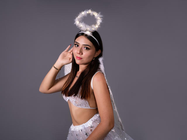 AlannaJonson with dark hair, wearing a halo, white lace bra, and sheer veil, pointing at her face with a slight smile, showing her small tits and slim body.