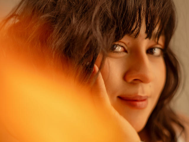 JuliettaHott's face with brown wavy hair, soft smile, and glowing eyes, partially obscured by a warm orange blur on the left. Her cute nose and slightly parted lips are visible.