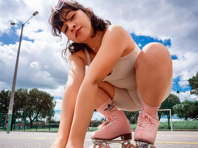 JuliettaHott crouches down in pink roller skates, wearing a ribbed beige tank top and shorts, with her ass up and tits barely visible, under a cloudy sky.