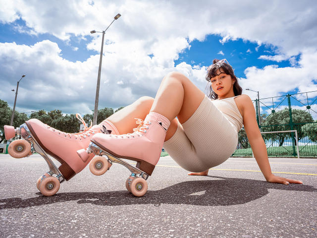 JuliettaHott with pink roller skates, white ribbed dress, and cute pink socks, sitting on asphalt, legs bent, looking at camera, cloudy blue sky, trees in background, sexy.