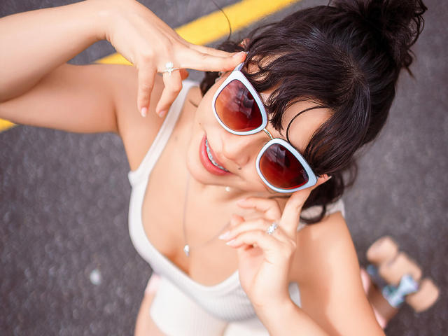 JuliettaHott with big tits, wearing white sunglasses, adjusting them with hands, wearing a white tank top, black hair in a bun, yellow line, and a ring on her finger.