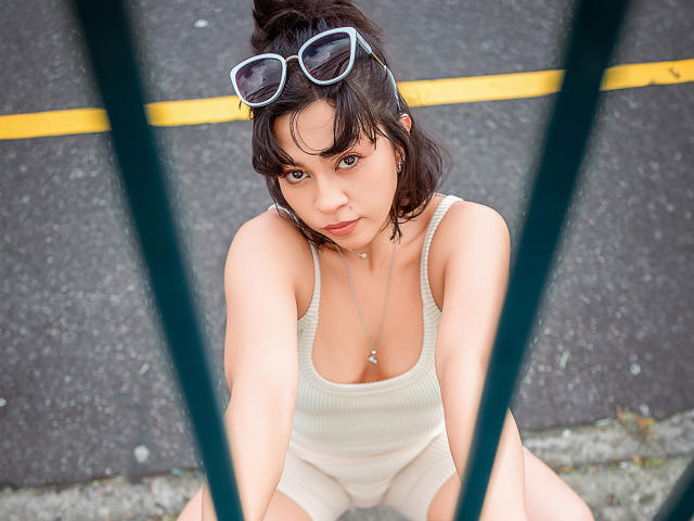 JuliettaHott with dark hair in a messy bun, wearing a beige ribbed tank top, sitting with legs apart, white sunglasses on head, looking up, yellow line in background.