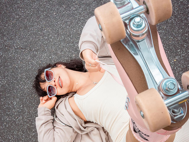 JuliettaHott lying on her back with a skateboard, wearing a white tank top, grey hoodie, and pink glasses, looking up with a smile, her tits partially visible.