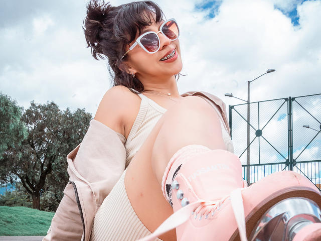 JuliettaHott with pink roller skates, white sunglasses, and a beige dress, showing her ass and a smile, jacket off, in a sunny outdoor setting. Her hair is in a messy bun.