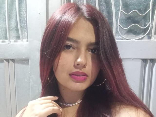 MiiaFoxiy with long, red-purple hair, pink lipstick, and hoop earrings, wearing a bead necklace, has a cute face and a slight, confident smile.