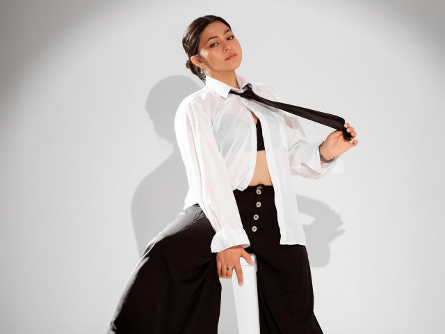 JuliettaHott with dark hair, white shirt, black tie, black high-waisted pants, and black bra, holding tie, showing her tits, shadow on white background.