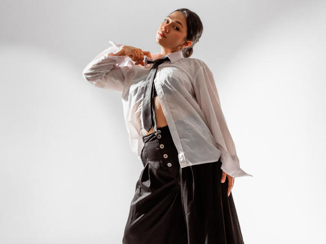 JuliettaHott with a white shirt, black tie, and high-waisted black skirt, buttoning her shirt, exposing her tits, with her hand on her chin, in a studio.