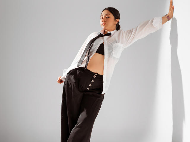 JuliettaHott has black tits, wearing a white shirt, black tie, and high-waisted black pants with buttons, leaning against a wall, with her arm raised and a slight smile.