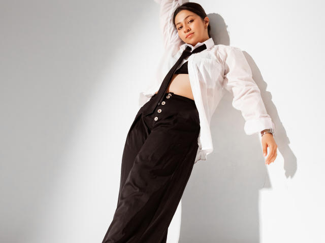 JuliettaHott with a slim, sexy build, wearing an open white shirt, black pants, and a black tie, exposing her tits, leaning against a white wall, looking down.