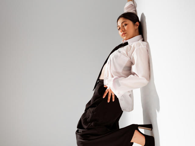 JuliettaHott with a white shirt, black dress, and black heels, leaning against a white wall, hand on hip, looking seductive, black headband, soft light, confident pose.