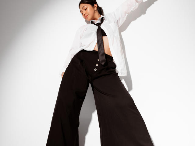 JuliettaHott with her tits exposed under a white shirt, black tie, and high-waisted black pants, arms raised, standing in a low-angle shot, looking down with a relaxed expression.