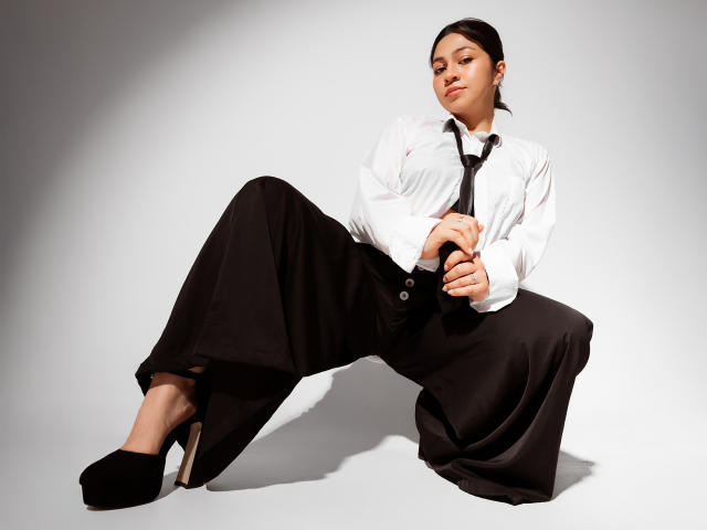 JuliettaHott, wearing a white shirt and black tie, sits with legs spread, black high heels, and black pants, looking confident with hands near her knees, in a studio.