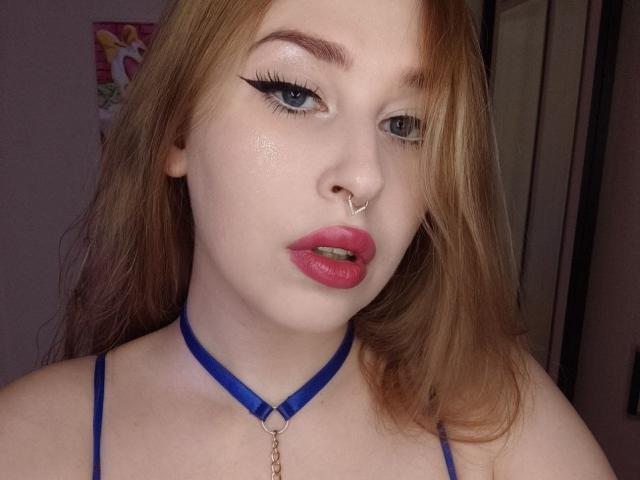 AliceLovels with red hair, blue choker, nose ring, and pink lipstick, showing off her tits with thick eyeliner and slightly parted pink lips, looking at the camera.
