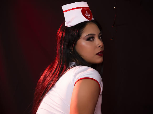 AnnieHill in a nurse costume with a white hat and red cross, looking over her shoulder, showing her dark, shiny hair, and red lipstick, with her tits visible.