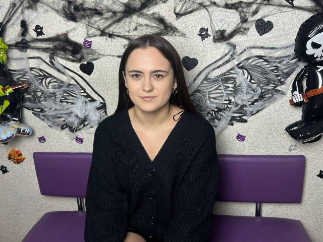 MarySweetSuprem has long brown hair, fair skin, and is wearing a black cardigan. She's sitting on a purple bench with a smoky, abstract wall behind her.