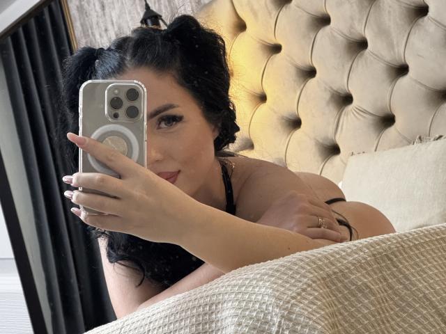 BellaaNoir69 with puffy tits, black curly pigtails, holding a phone, taking a selfie, lying on a bed, wearing a black strap, tan quilted headboard in the background.