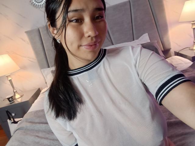 Karollteyes with shiny tits, wearing a sheer white shirt with black stripes, has long black hair in a ponytail, smiling at the camera in a bedroom.