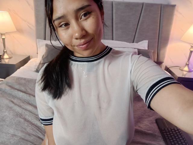 Karollteyes in a sheer white tee with black stripes, wet chest showing small tits, standing in a bedroom with a bed and lamps. She's smiling, dark hair in a ponytail.