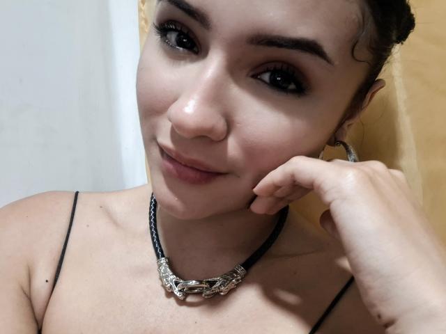 LizzeJoy with dark brown eyes, curly hair, and a subtle smile, wearing a black necklace with a silver bone pendant, touching her cheek with her right hand.