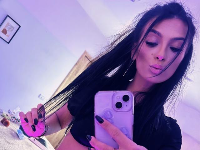 KinkyAmelie mit langen schwarzen Haaren, voller Lippen, schwarzen Nägeln, einem schwarzen Oberteil, die ein pinkfarben beleuchtetes Handy hält, ein Selfie macht und ihre kleinen Brüste zur Schau stellt, in einem violett beleuchteten Raum..