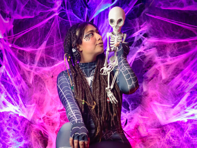 BeckyHuston with braided hair, wearing a Spider-Man suit, holding a skeleton, pink and purple neon spiderwebs, and face paint on her cheek, looking at the skeleton with a smirk.