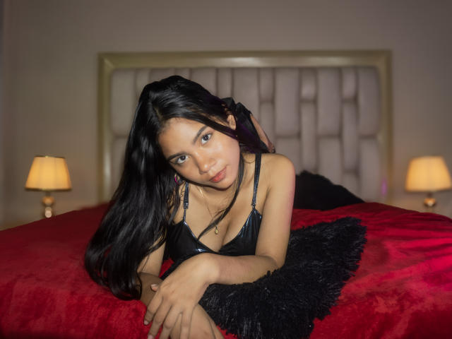 NatashaRuiz with long black hair, wearing a black leather bra, leaning on a red bed with black fur pillow, looking at camera, soft bedroom light, slight smile.