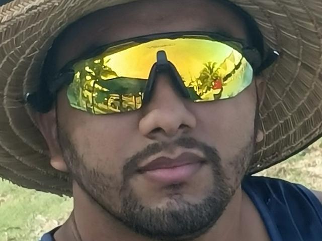MarooHot with a dark beard, wearing yellow reflective sunglasses and a straw hat, has a subtle smile and a dark shirt, showing a reflective palm tree in the lenses.