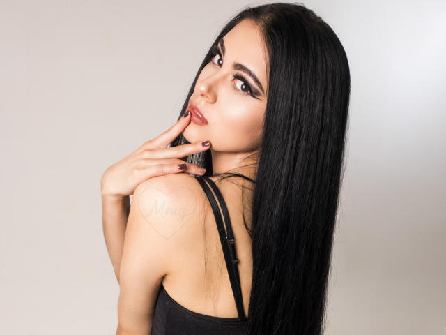 AmberLenox with long black hair, looking over shoulder, biting finger, showing tattoo on shoulder, wearing black top, pale skin, red lipstick, and dark nail polish.