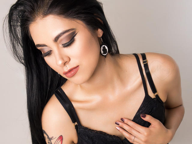 AmberLenox with long black hair, closed eyes, and makeup, wearing a black lace bra, holding her tits, shiny skin, ring nose, tattoo on her right arm, and hoop earrings.