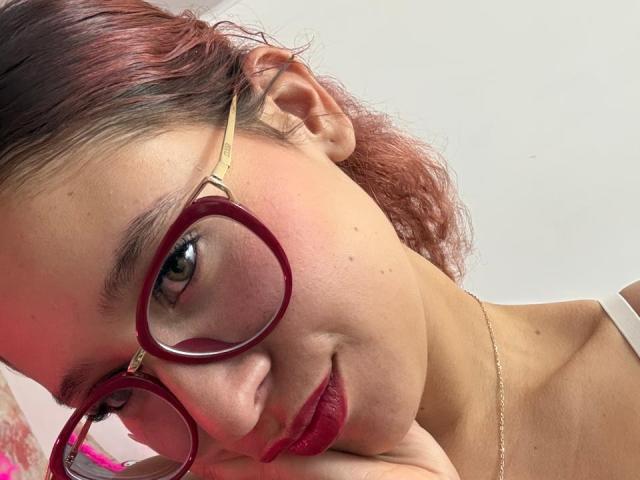ValeryScot porte des lunettes rouges, des cheveux bouclés roses et du rouge à lèvres rouge, et regarde vers le bas avec un léger sourire. Ses fesses sont légèrement visibles dans le coin supérieur gauche..