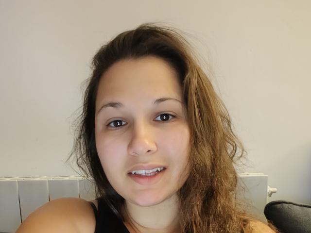 MinetteSexy69 has wavy brown hair, brown eyes, and a slight smile showing teeth. She's wearing a black top, with a white radiator in the background.