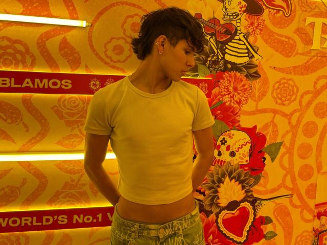 Angelitocute with short dark hair, wearing a tight yellow shirt and faded jeans, standing with hands on hips, showcasing a slim, cute butt. Bright, colorful background.