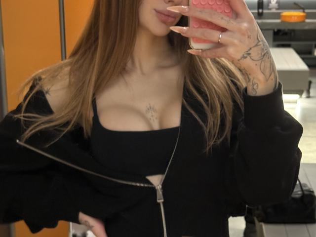 SensualMila mit braunen Haaren, schwarzem Top, das ihre Brüste zeigt, schwarzer Jacke mit Reißverschluss, Tattoos an den Händen, macht ein Selfie mit einem rosa Handy, in einem schwach beleuchteten Raum..
