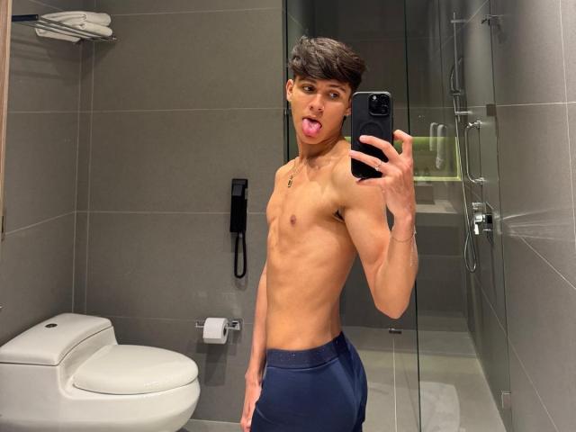 Angelitocute, shirtless with a slim, toned body, sticking out his tongue, taking a mirror selfie in a bathroom, wearing dark blue shorts, holding a black smartphone.