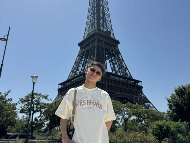Angelitocute with sunglasses, wearing a white Westford t-shirt, standing in front of the Eiffel Tower, hands in pockets, bag on shoulder, smiling. Clear blue sky.