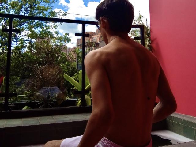 Shirtless Angelitocute with a toned back and small tattoo on the right side, sitting on a green-tiled balcony, wearing white and pink striped shorts, looking outside.