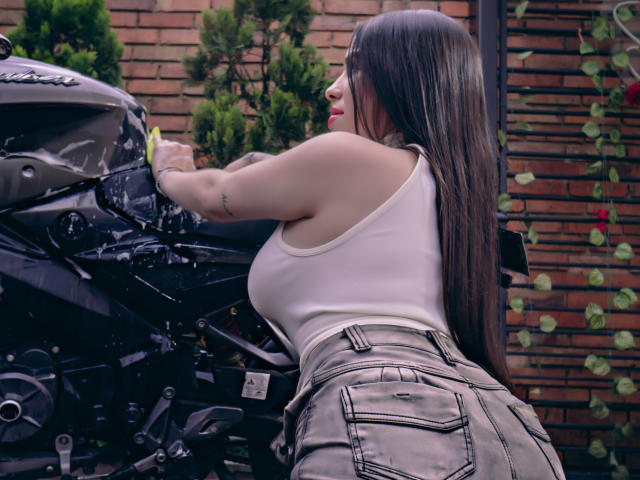 SteffaCandleX with long brown hair, white tank top, and gray jeans, leaning on a black motorcycle, washing it, showing her ass, tattoo on arm, brick wall background.