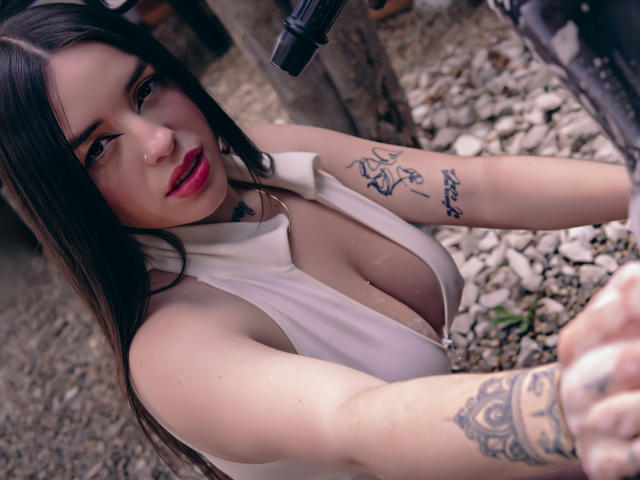 SteffaCandleX with long black hair, pink lipstick, white sleeveless top, cleavage, tattoos on arm and hand, looking at camera, gravel ground, black camera above.