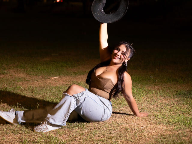 LorensDouce with tits in a brown crop top, holding a black hat, sitting on grass, wearing light blue jeans, and white shoes, smiling, arm raised, dark background.