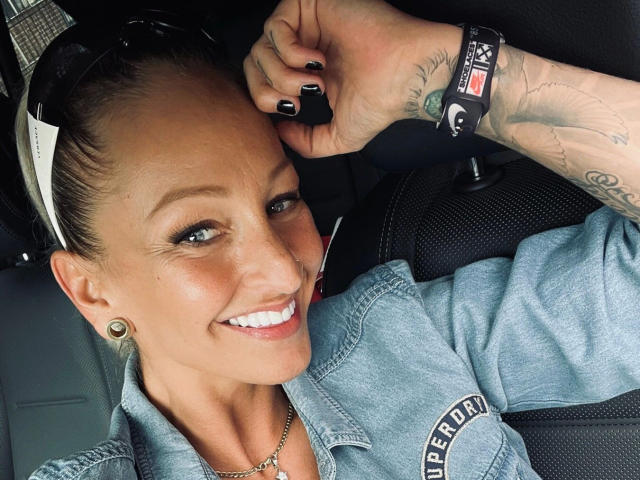 AlphaMaedchen-hot with tattoos on her arm, black nail polish, blue eyes, and a smile, wearing a denim shirt, holding a black object, and sunglasses on her head.