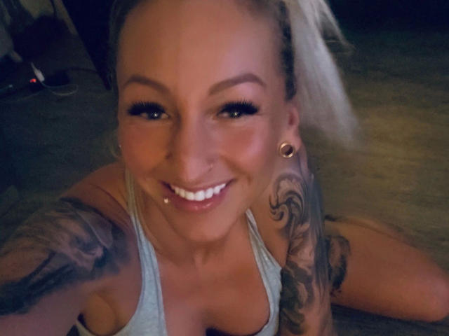 AlphaMaedchen-hot with blonde hair, large tits, tattoos on arm, smiling, nose ring, lip piercing, hoop earrings, wearing gray tank top, taking selfie from above.