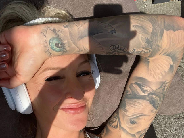 AlphaMaedchen-hot lying down, arm covering eyes, white headphones, tattoos on arm, smiling, sunlit, blonde hair, white skin, tattoos include eye, bird, and face.