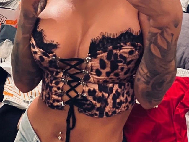 AlphaMaedchen-hot's taut, tan-skinned torso in a leopard-print, lace-up, black-fringed bustier, showing off her boobs, tattoos on her arms, belly button piercing, and light blue jeans.