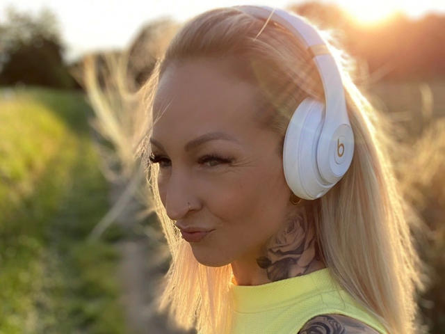 AlphaMaedchen-hot with blonde hair, white headphones, and a yellow top, has a tattoo on her neck and a small nose ring, looking sideways with a slight smile.