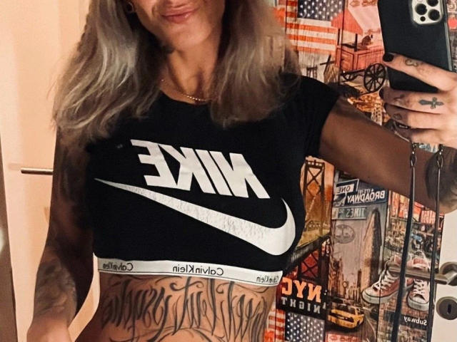 AlphaMaedchen-hot with grey hair, black Nike crop top showing tits, white Nike logo, and tattoos on her belly. She's holding a phone, smiling, in a mirror selfie.