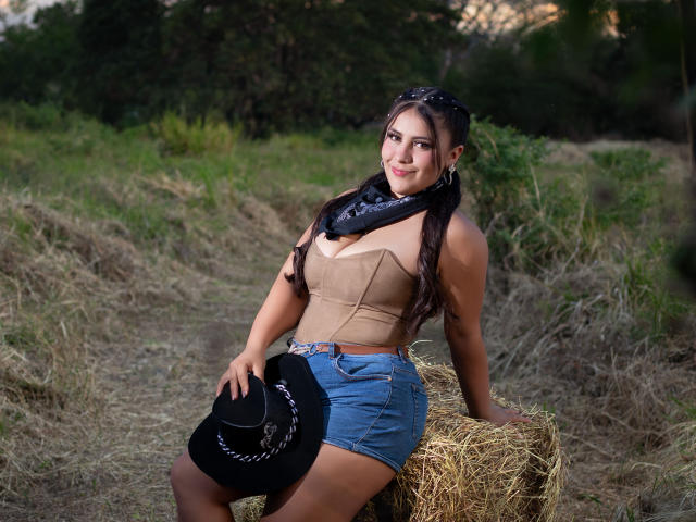 LorensDouce with big tits, sitting on hay, wearing a tan top, blue shorts, black scarf, holding a black hat, outdoors, evening light, long dark hair, slight smile.