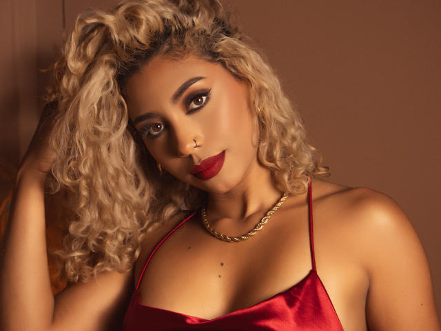 LilithWalker with curly blonde hair, red satin top, gold chain, and red lipstick, has a nose ring and sexy brown eyes, showing off her tits. Warm brown background.