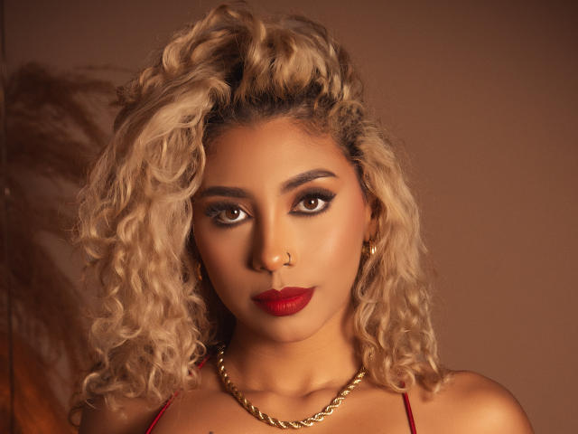 LilithWalker has curly blonde hair, dark eyebrows, red lipstick, and a gold chain necklace. She's wearing red straps and gold earrings, looking directly at the camera.